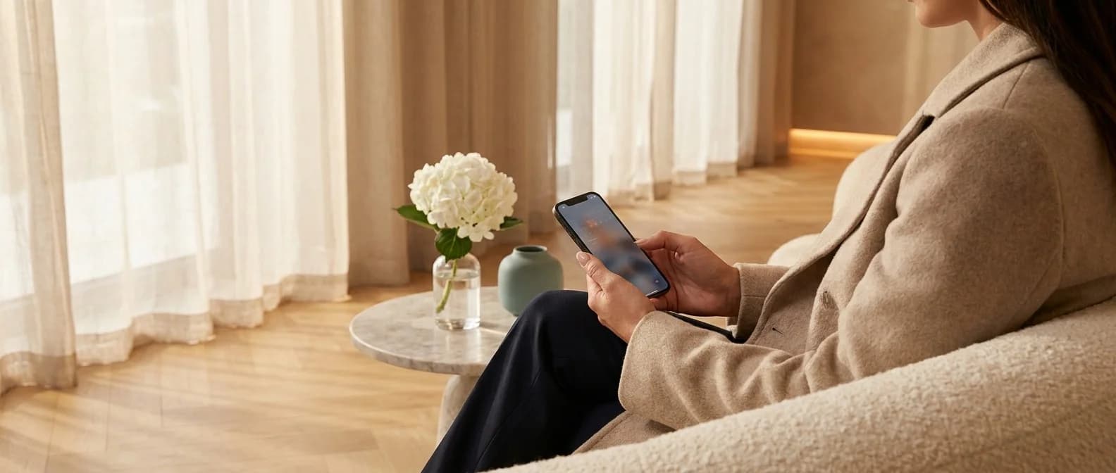 Patientin auf cremefarbenem Bouclé-Sofa im warmen Wartebereich der Klinik bucht ihren Termin am iPhone