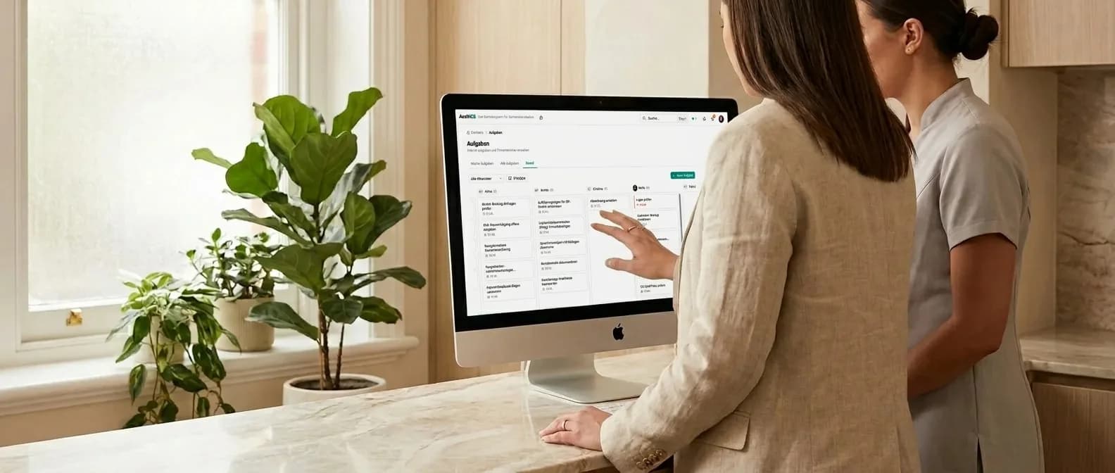 Praxismanagerin und MFA stehen am Marmor-Empfangstresen vor einem 27-Zoll-iMac, der das AesthOS-Aufgaben-Board mit Mitarbeiter-Spalten und Aufgabenkarten zeigt